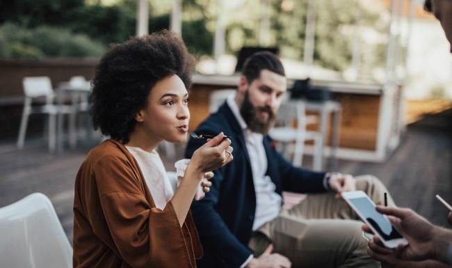 A beautiful community at the heart of all your passions woman sits and eats outside while someone holding a cell talks to her